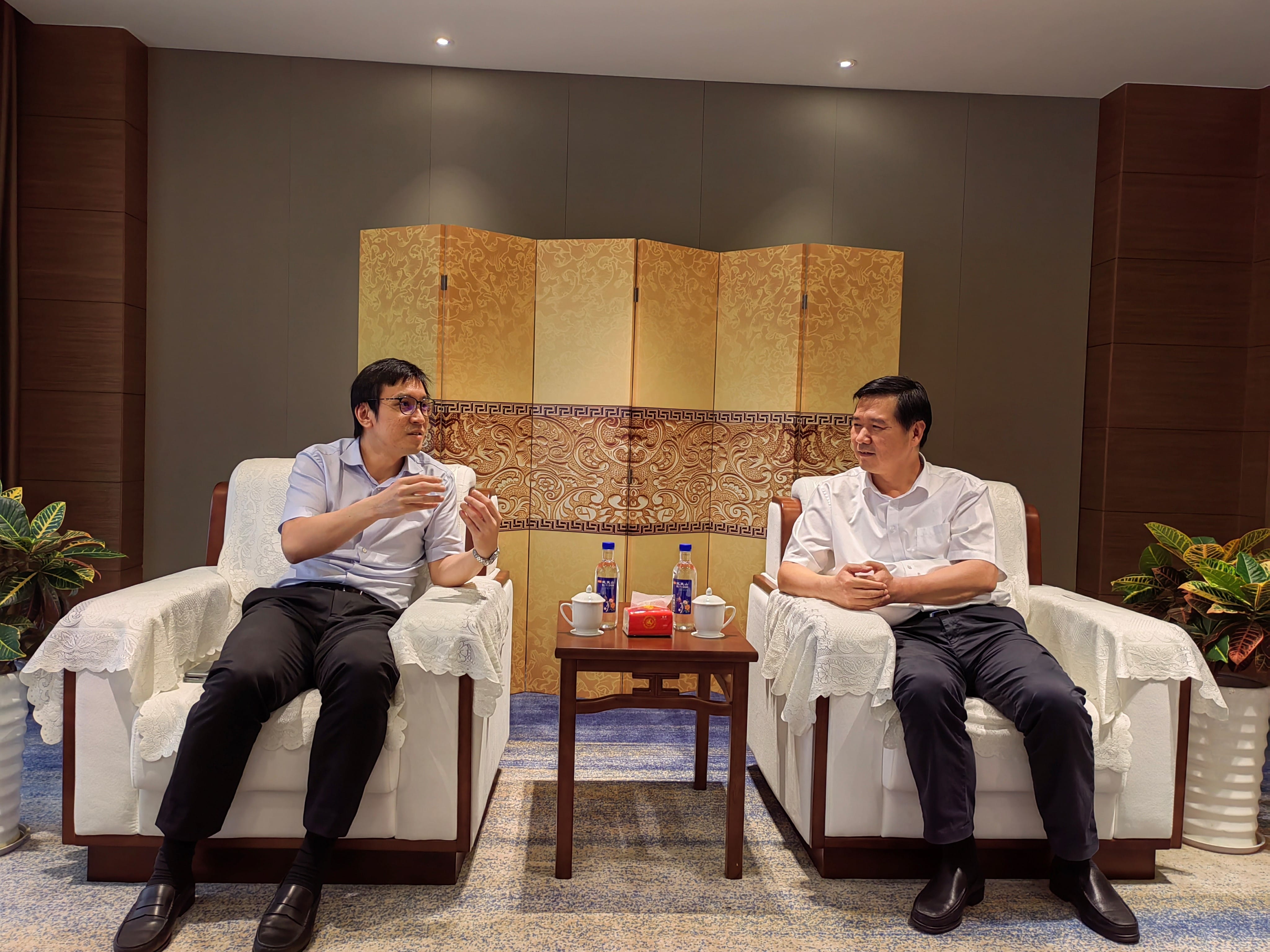 Two men in shirts sit in white armchairs, facing each other in a formal setting. Bottled water on a table between them.